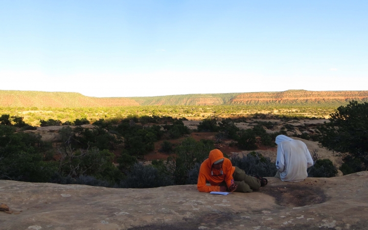 Two people sit on a rock overlooking a desert landscape and journal.